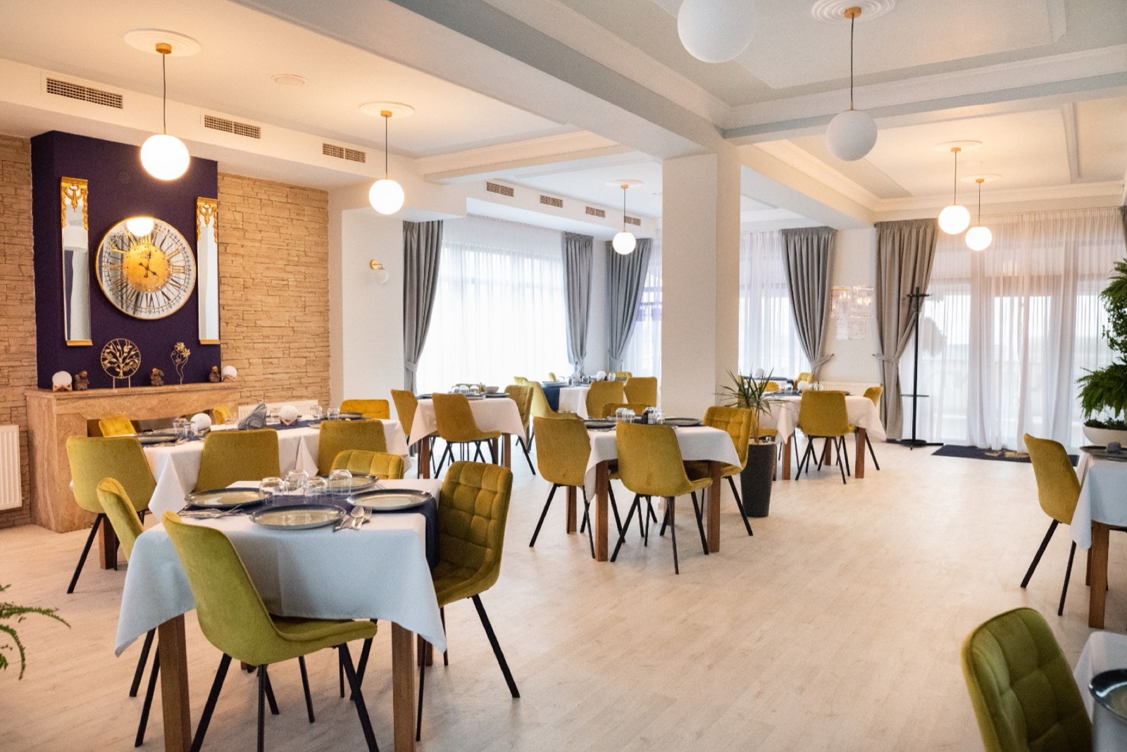 Salonul principal al restaurantului, cu mese așezate, scaune de catifea muștar și peretele indigo cu ceasul auriu